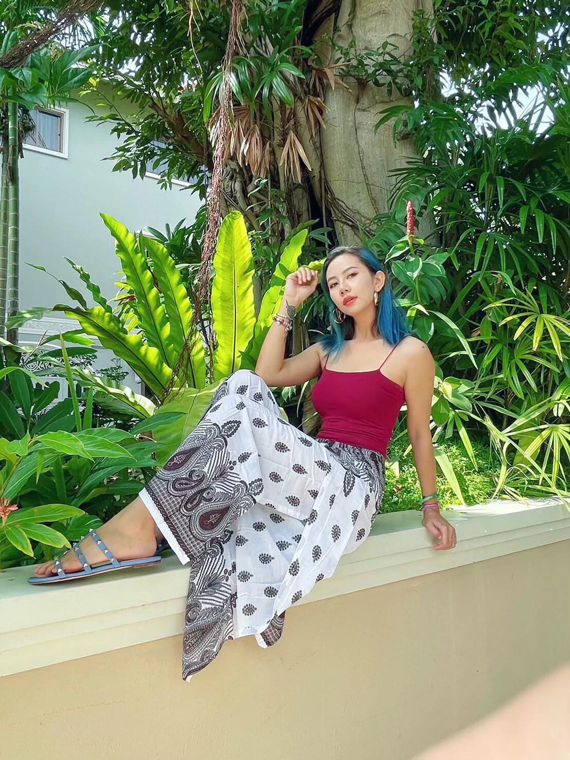 A beautiful lady is sitting on a ledge, surrounded by lush greenery, wearing boho wrap pants with a distinct pattern and a red top