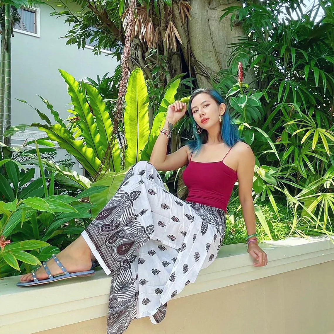 A beautiful lady is sitting on a ledge, surrounded by lush greenery, wearing boho wrap pants with a distinct pattern and a red top