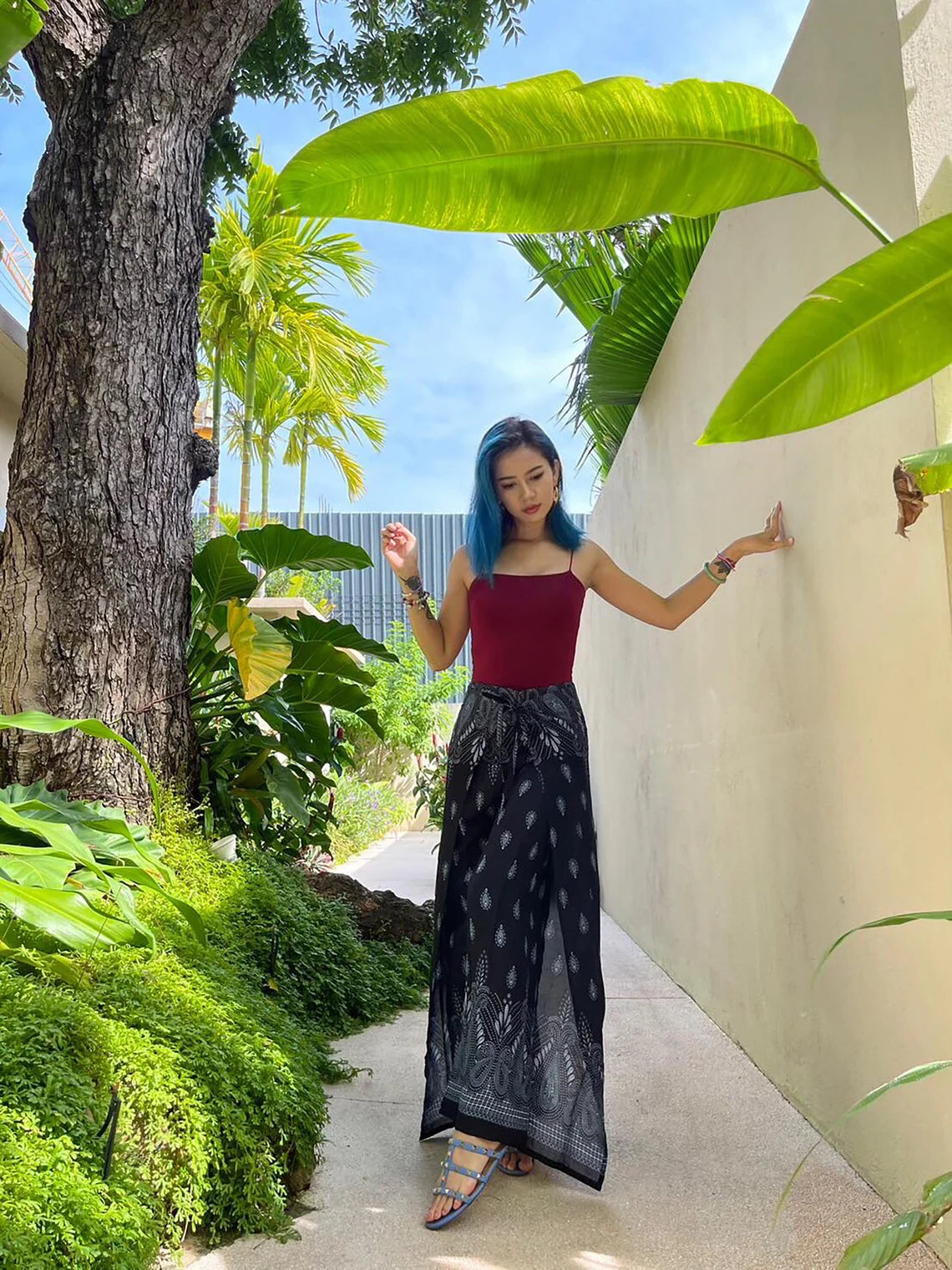 A person wearing boho black wrap pants paired with a red top, standing outdoors surrounded by greenery and a white wall