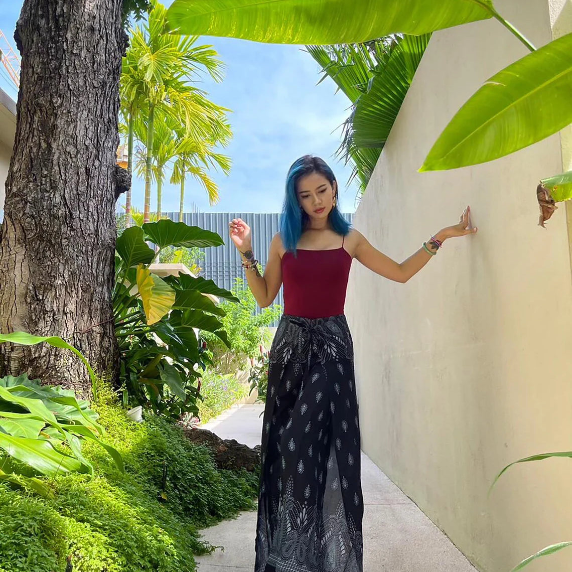 A person wearing boho black wrap pants paired with a red top, standing outdoors surrounded by greenery and a white wall