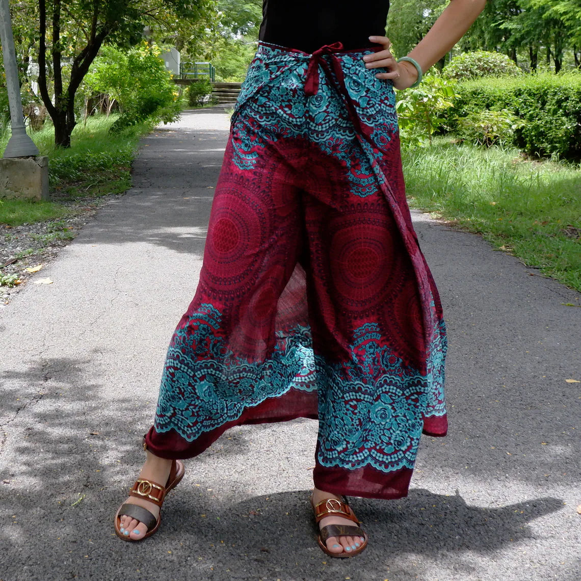 Vibrant boho beach wrap pants with teal and burgundy patterns, black top, and brown sandals in a lush outdoor setting.