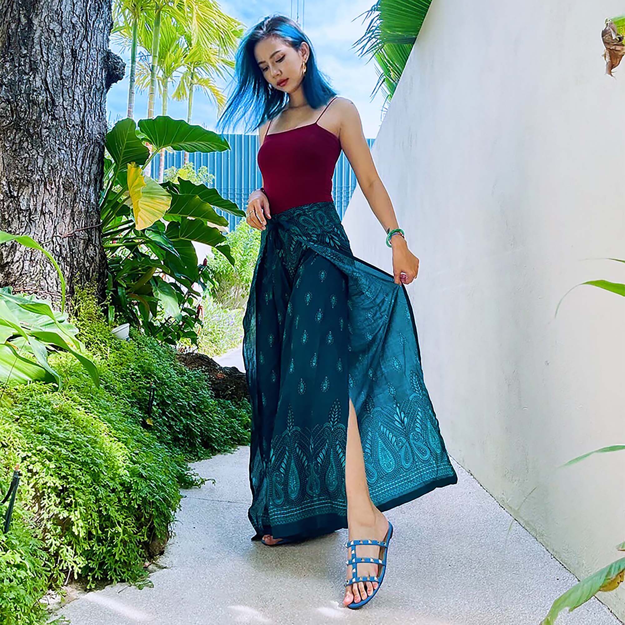 Woman in teal boho wrap pants walking in tropical garden with trees and white wall background

