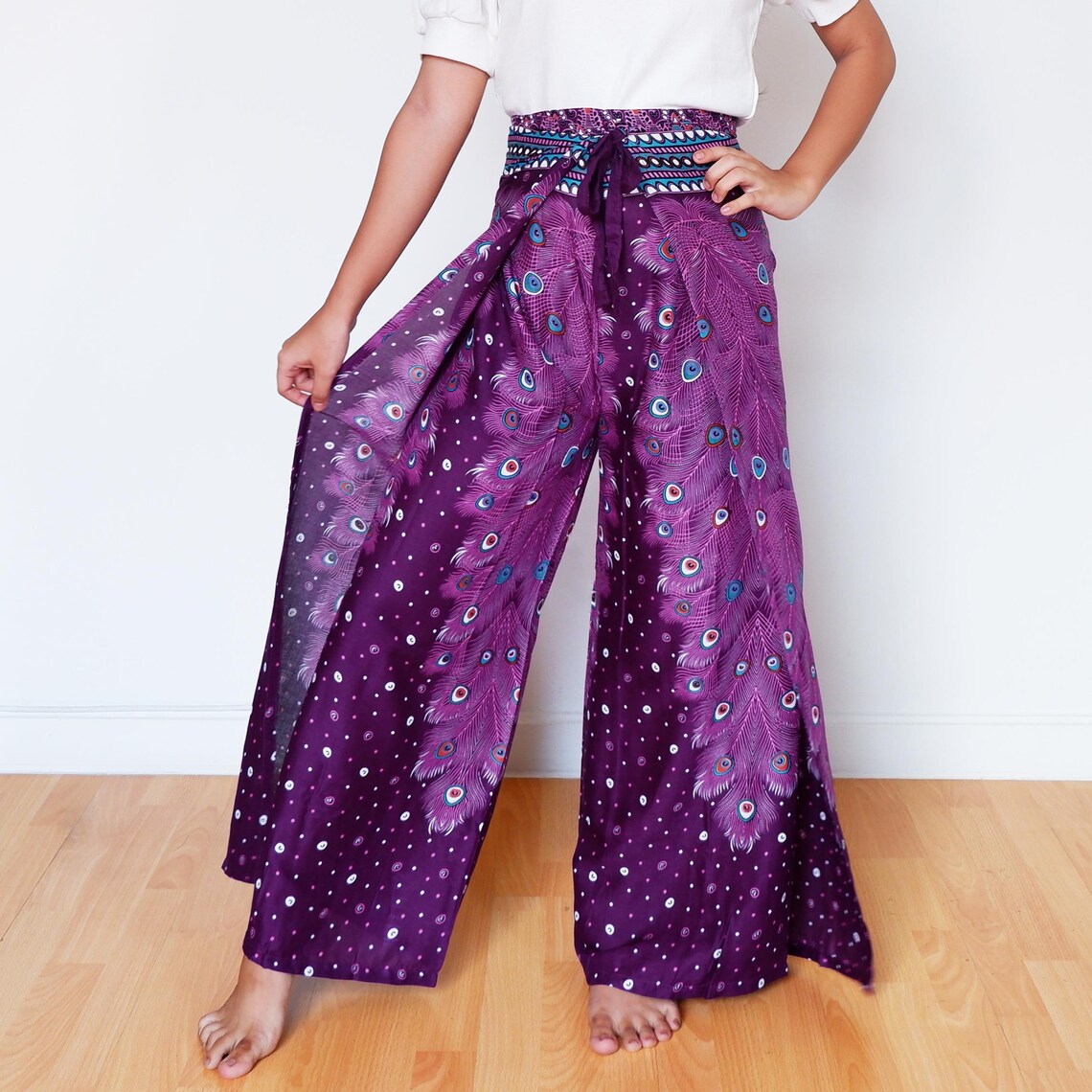 Purple boho wrap pants with handmade peacock design, model holding open leg panel
