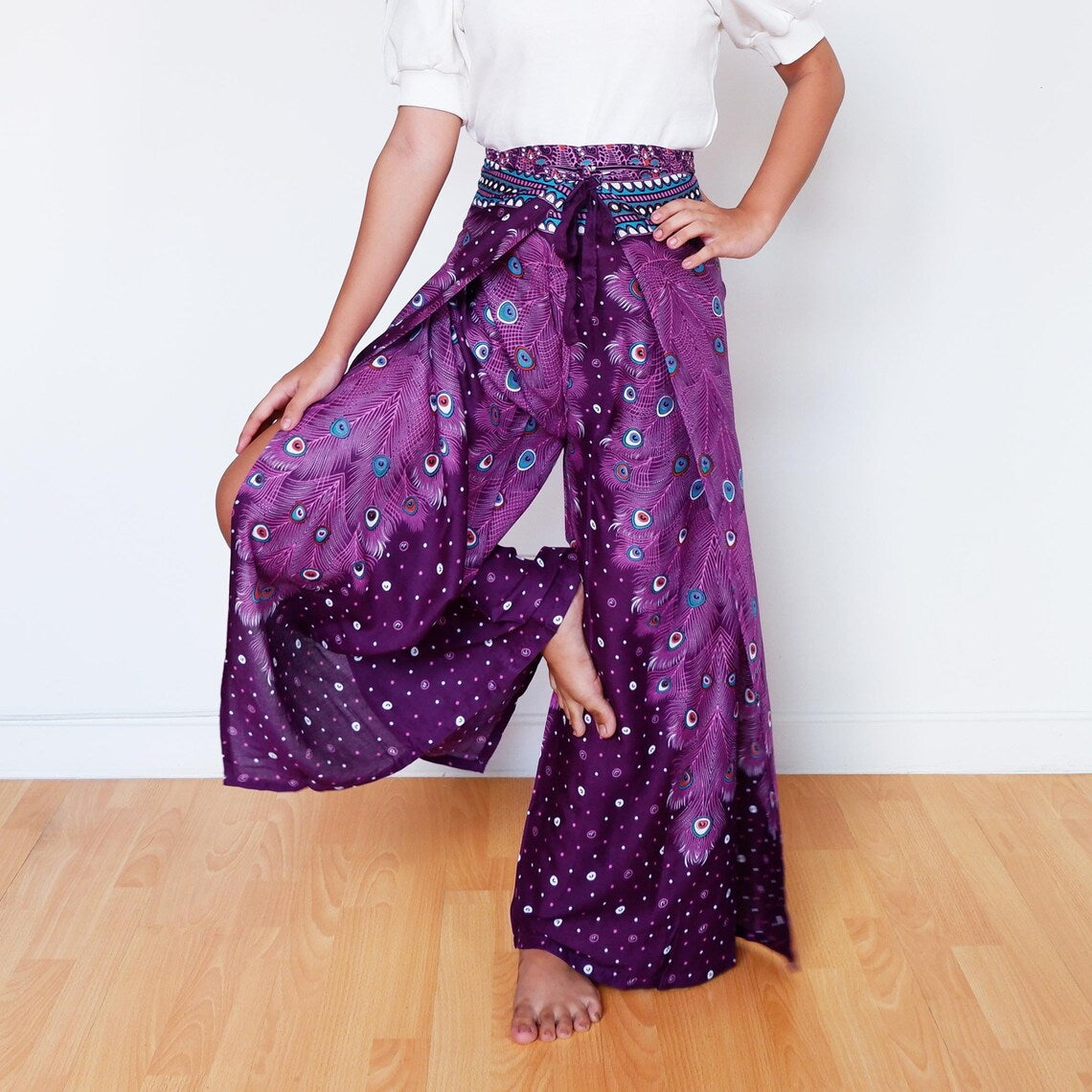 Model playfully lifting leg in purple wrap pants with peacock feather print

